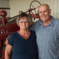 Charlotte and her husband, an older man in a blue button-up shirt, posing for a photo in front of an older red tractor.