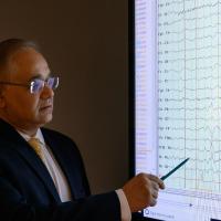 Dr. Siddharth Kapoor, pictured in a dark room looking at a projector screen displaying monitor scans while he points at the screen with a pen.