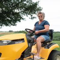 A photo of Charlotte smiling at the camera as she drives a yellow riding lawn mower.