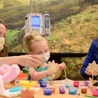 A candid photo of Christina Harper, a young patient, and her mother playing with wooden flowers. Christina is a white woman with long light brown hair. She is wearing dark blue scrubs and a light blue face mask.