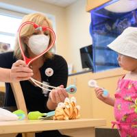 A candid photo of Jennifer holding a heart-themed toy stethoscope in front of a young female patient. The patient is a young white girl in a white bucket hat and a pink and yellow strappy top.