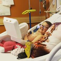 An older photo of Ellie laying in her hospital bed, enjoying a snack and giving the camera a cheesy smile.