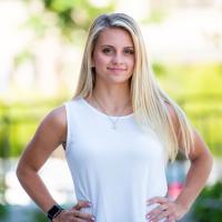 Emma, wearing a white tank top, stands in an outdoor setting with her hands on her hips. She’s smiling and looks determined and confident.