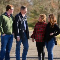 Fielden and her dad, Doug, smile at each other. They are on their family farm. Doug, an older white man with graying hair, is wearing a white collared shirt and a navy jacket. Fielden is wearing a black turtleneck sweater.