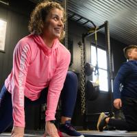 Geri smiles as she works out in the gym next to her son. She is doing a lunge and looking off-camera.