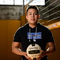 Gil holds a volleyball in front of a volleyball net in a gym.