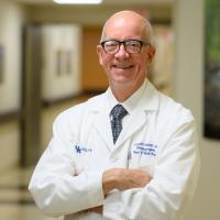 Dr. Joseph Valentino, an older white man with short white hair, smiles for the camera with his arms crossed. He is wearing dark-rimmed glasses, and a white lab coat over a white button-up shirt with a navy tie.