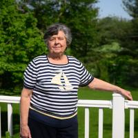 A photo of Helen resting her hand on the railing of her back porch as she looks into the camera and smiles.