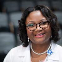 Joy smiles brightly while wearing her white nurse practitioner's coat in a lecture hall.