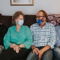 Mae, wearing a turquoise shirt and matching earrings, sits on the couch with three middle-aged men. Each of them are wearing a facemask and are looking at one another.