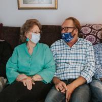 Mae, wearing a turquoise shirt and matching earrings, sits on the couch with three middle-aged men. Each of them are wearing a facemask and are looking at one another.