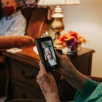 A close-up of Mae wearing a mask at home, on a video call with a young white woman who is smiling widely.