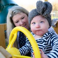 MaKenzie plays with a steering wheel on a playground jungle gym. Her parents are supporting her.