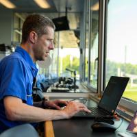 A candid photo of Matt working on his laptop while sitting in a box that overlooks the field and stadium.