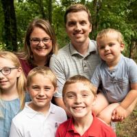 A group shot of Max and his family, standing together in an outdoor setting. He’s surrounded by his mom and dad, his blonde-haired older sister, his blonde twin brother. His dad is holding Max’s blonde-haired toddler brother.