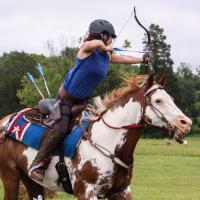 Miko rides a running horse, who is running toward the camera. Miko is turned away from us, getting ready to shoot an arrow at a target off-screen.