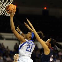 An action shot of Ogechi during a game, jumping up to the basket with a basketball in her hand and an opposition defender behind her.