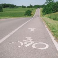 Close up of the biking lane symbol painted on the road Patty likes to bike on. The road is surrounded by beautiful, grassy rolling hills.