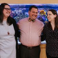 Dr. D’Orazio smiles and poses with Dr. Amanda Harrington and Terra Lucas. Terra is a young white woman with long, dark-brown hair. She is wearing a black cardigan over a gray shirt, and a necklace with a large brown gem on it. Dr. Harrington is a white woman with long brown hair. She is wearing a long-sleeve black shirt that has a pink and blue repeating pattern on it.