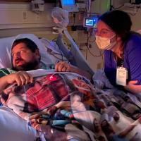 A candid photo of Ronald in his hospital bed looking at something off camera. A nurse is leaning over the railing of his bed and is also looking at something off camera.