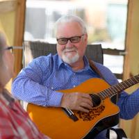 Ronnie plays the guitar as he sits on the porch with his wife Becky. She is an older white woman with long gray hair, and she is wearing a red and white checkered-pattern button-up shirt.