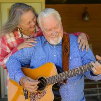 Becky smiles as she stands behind Ronnie. Her hands are on his shoulders as he plays the guitar in front of their house.