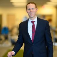 Dr. Austin Stone, a young-looking man with short brown hair, stands in the UK HealthCare orthopedic rehab facility. He is wearing a suit and tie and smiling.