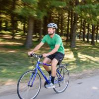 Ryan rides his bike along a road next to a clump of trees. The background is blurred slightly. He is wearing a green shirt and a helmet.
