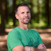 A portrait of Ryan in an outdoor setting. He has his arms folded in front of him and looks determined.