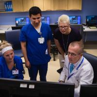 Dr. Day, dressed in a white doctor's coat and tie, sits in a chair as he looks at a computer with three of his co-workers sitting and standing next to him.