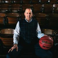 Todd takes a seat in some gymnasium seating and smiles with a basketball resting on his knee.