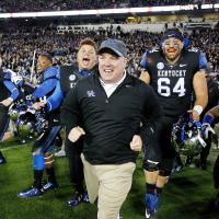 Grinning broadly, Coach Stoops runs out onto the field after a victory. He’s surrounded by extremely excited Kentucky football players.