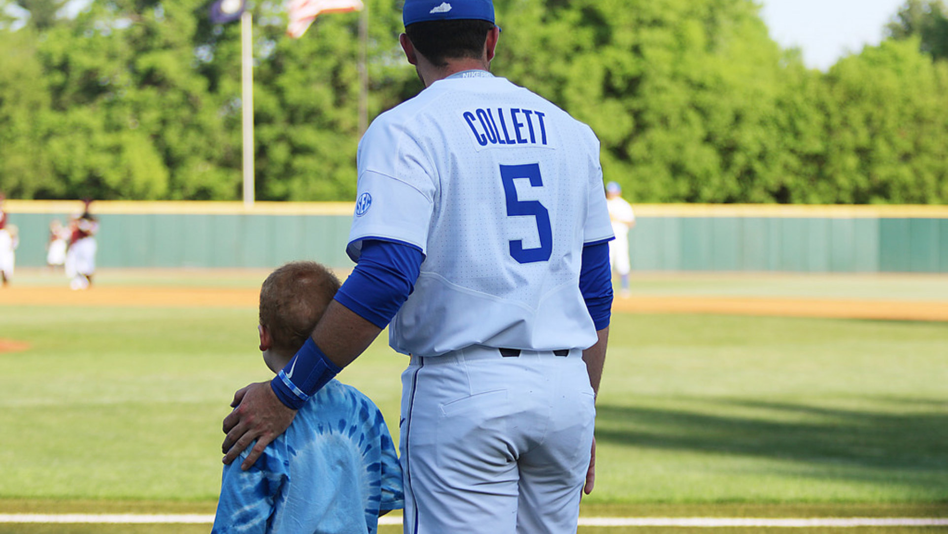 T.J. stands next to a young kid with short hair and a tie-dye shirt. The two are standing on the edge of a baseball field, watching other players warm up. T.J. has his hand on the kid’s shoulder.