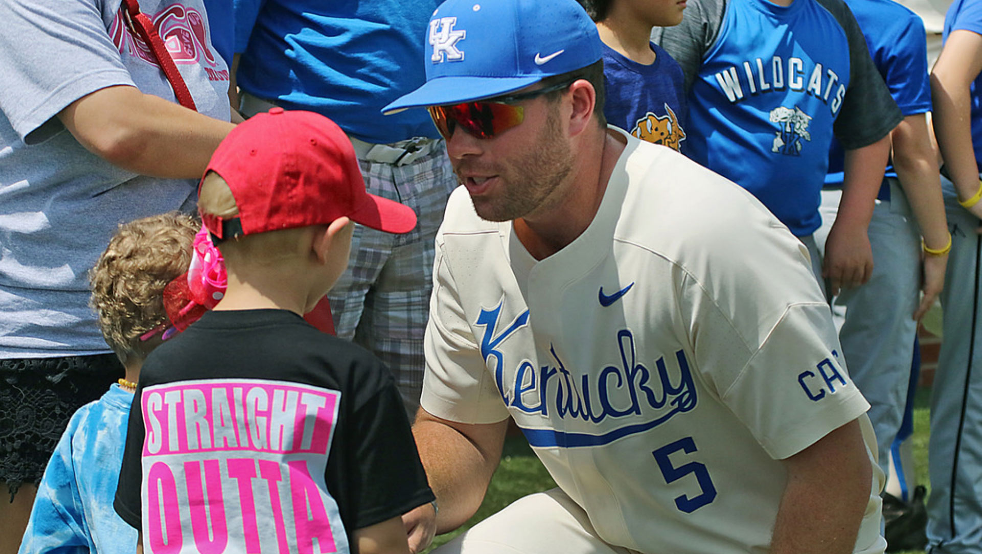 T.J. crouches down to talk to two kids. He has sunglasses and a baseball uniform on and there is a line of fans and baseball players behind him.