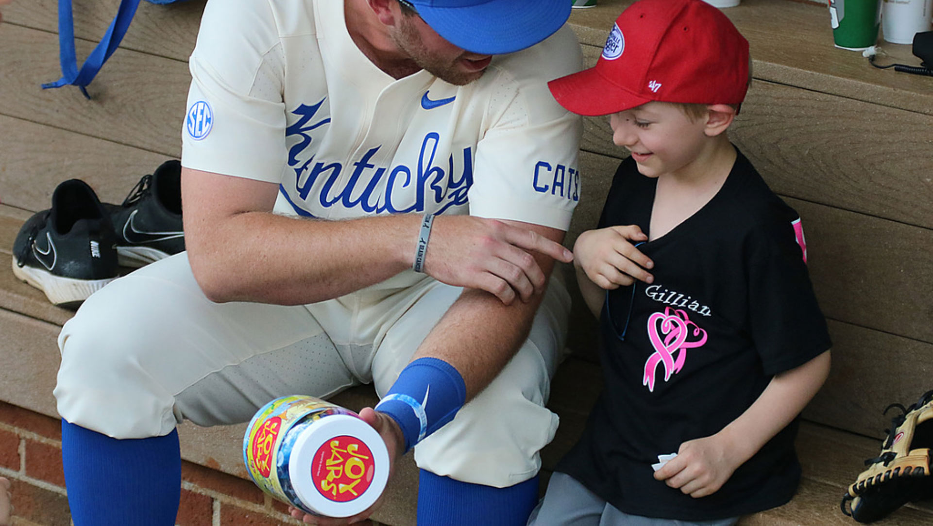 T.J. sits in the dugout with a young fan wearing aa baseball cap. T.J. is holding a JoyJar—a brightly colored plastic jar filled with toys—and is pointing at the kid playfully. Both are smiling.