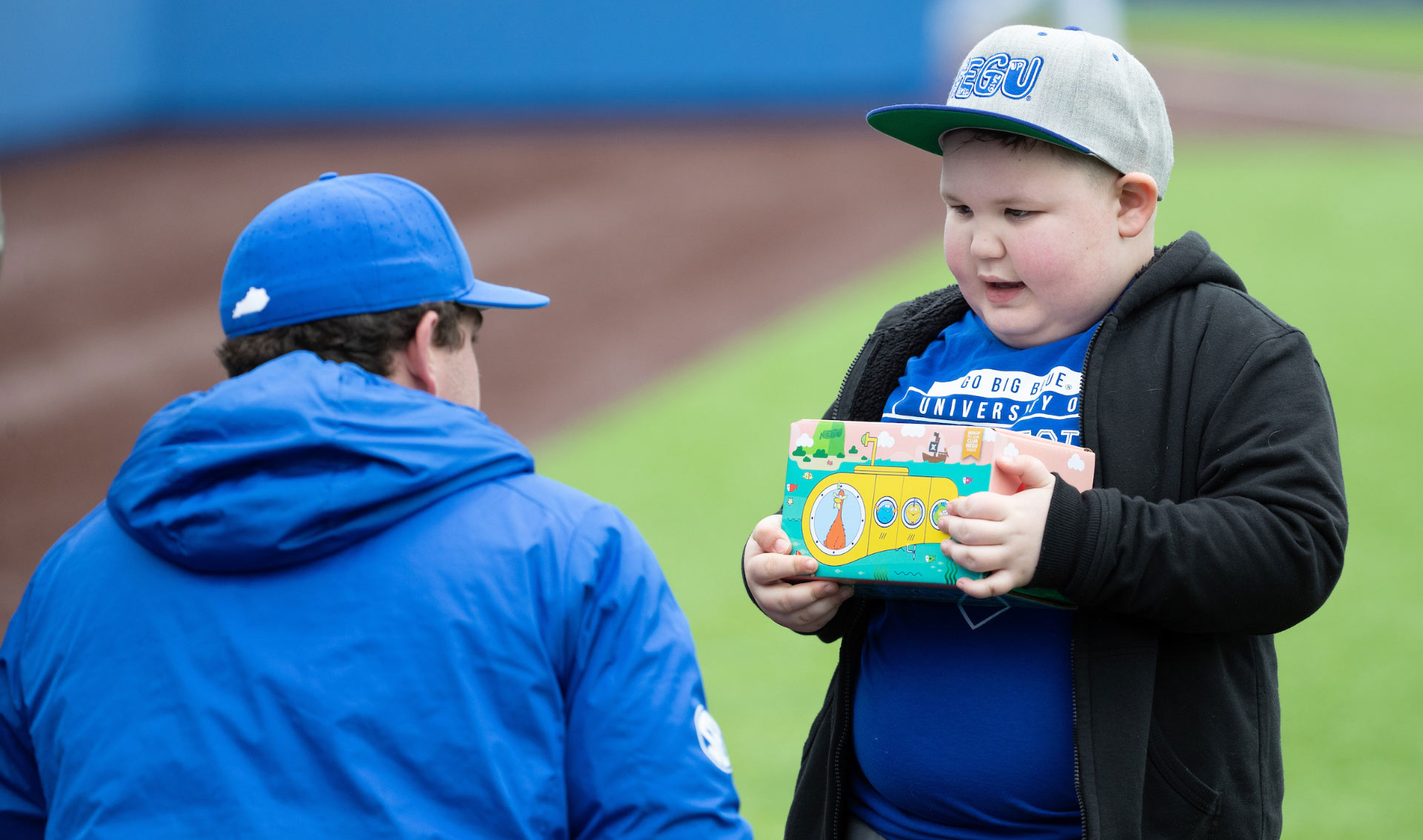T.J. crouches down to talk to a young kid wearing a baseball hat. The kid is holding a brightly-colored box. T.J. is wearing a baseball cap and a blue jacket.
