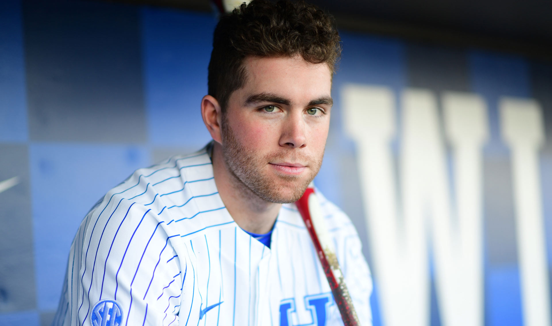 A portrait of T.J., with his face in focus and the background blurred. He is sitting in the dugout and has a baseball bat over his shoulder. He is wearing his baseball uniform and has a slight smile on his face.