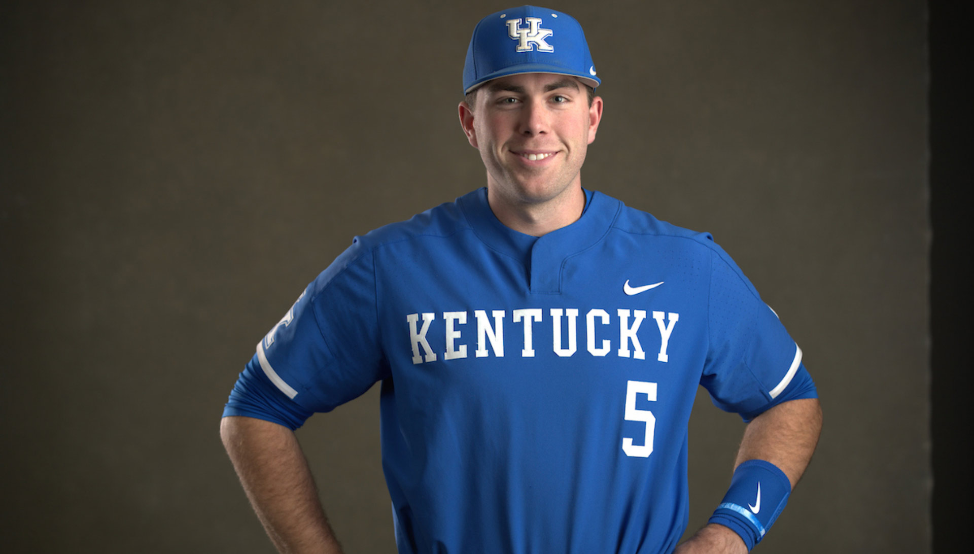 T.J. Collett, a young white man with brown hair, poses for a portrait against a gray backdrop. He has a Kentucky baseball uniform on, is standing with his hands on his hips, and is smiling.