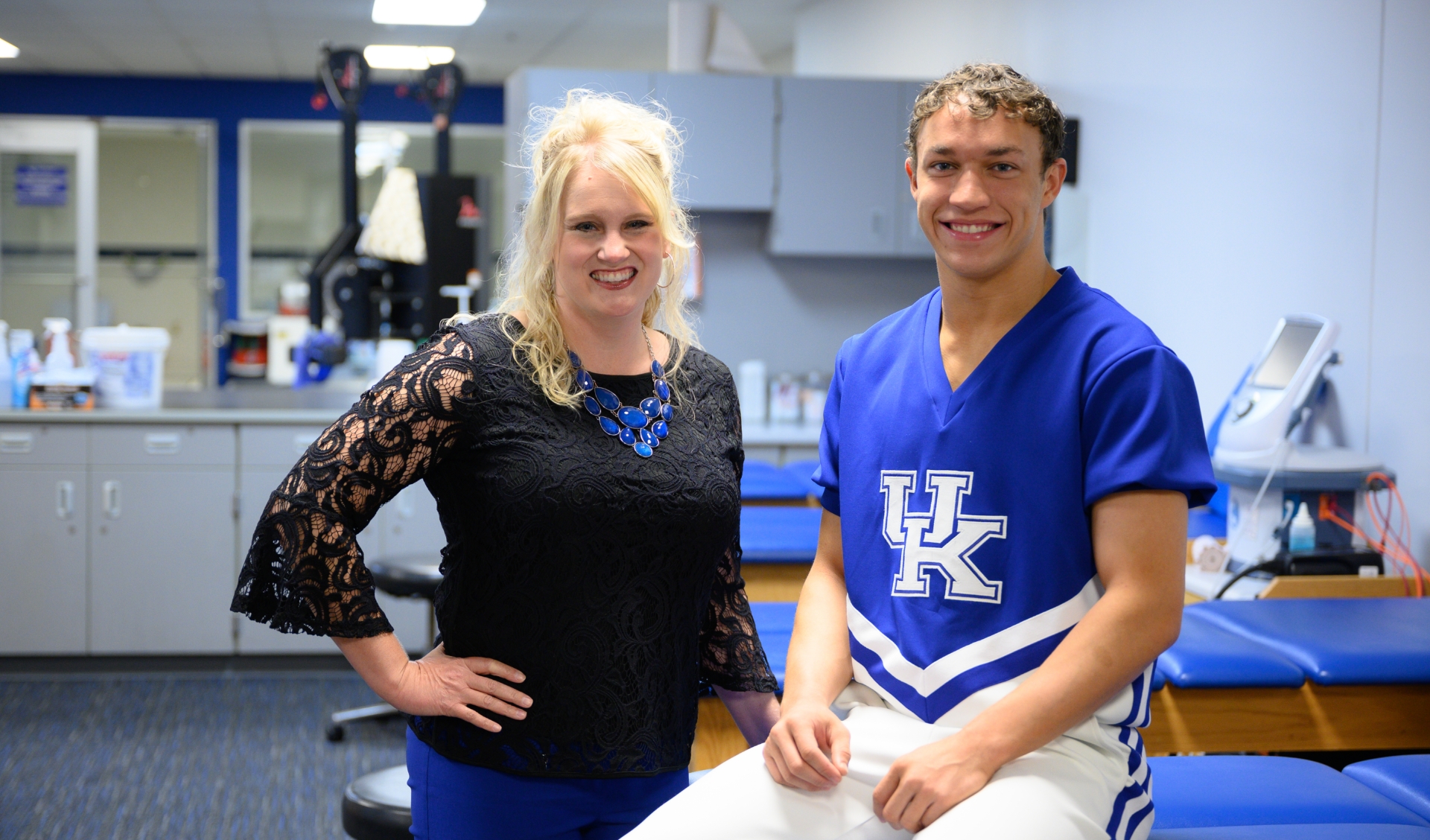 Travis smiles for the camera alongside Dr. Kimberly Kaiser, a white woman with long light-blonde hair. She is wearing a black, long-sleeve shirt with lace detailing and a large blue gemstone necklace.