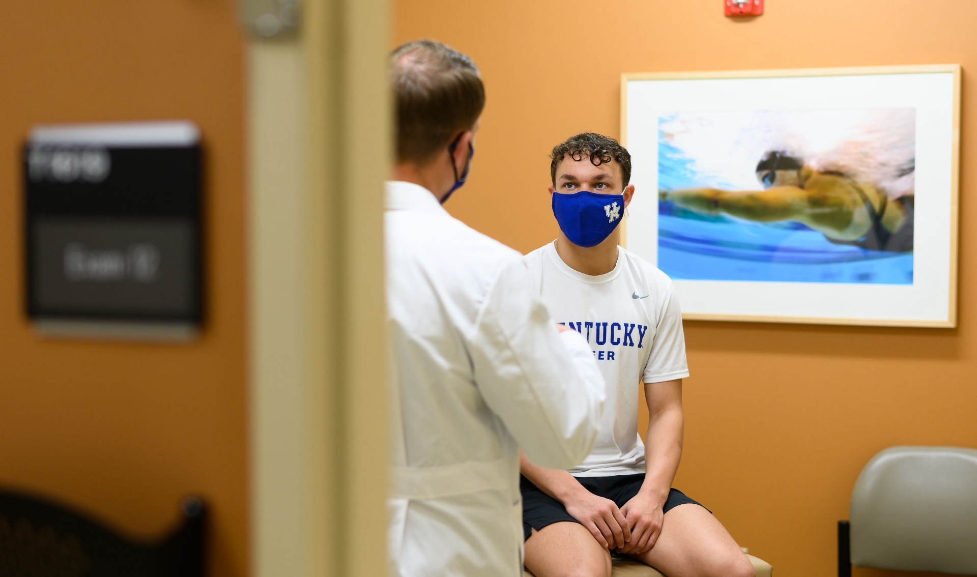 An over-the-shoulder shot of Travis having a conversation with Dr. Scott Mair. Dr. Mair is a white man with light brown wavy hair. He is wearing a white lab coat over blue scrubs, and a face mask.