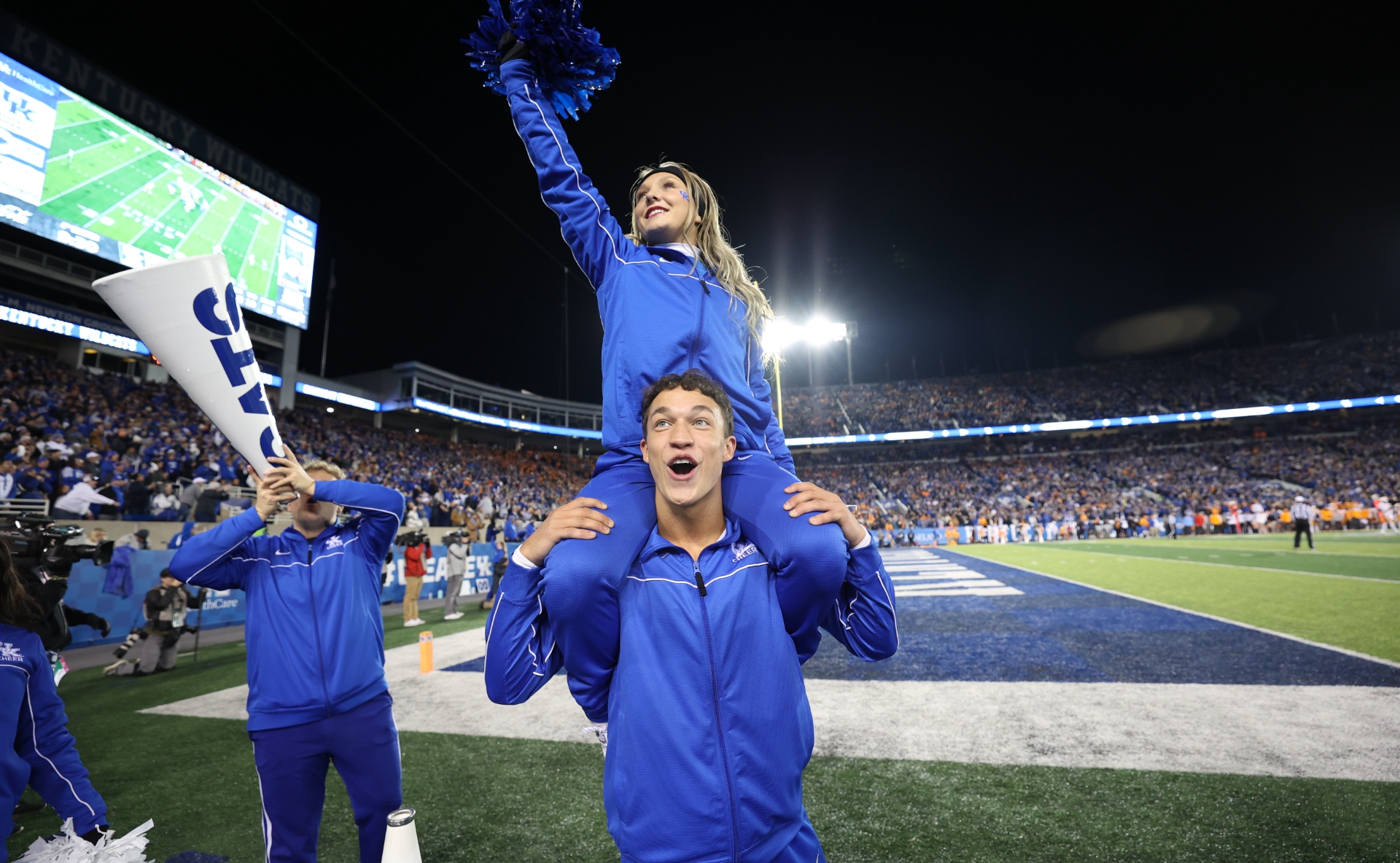 Travis cheers at a UK Football game, while a female cheerleader sits on his shoulders.