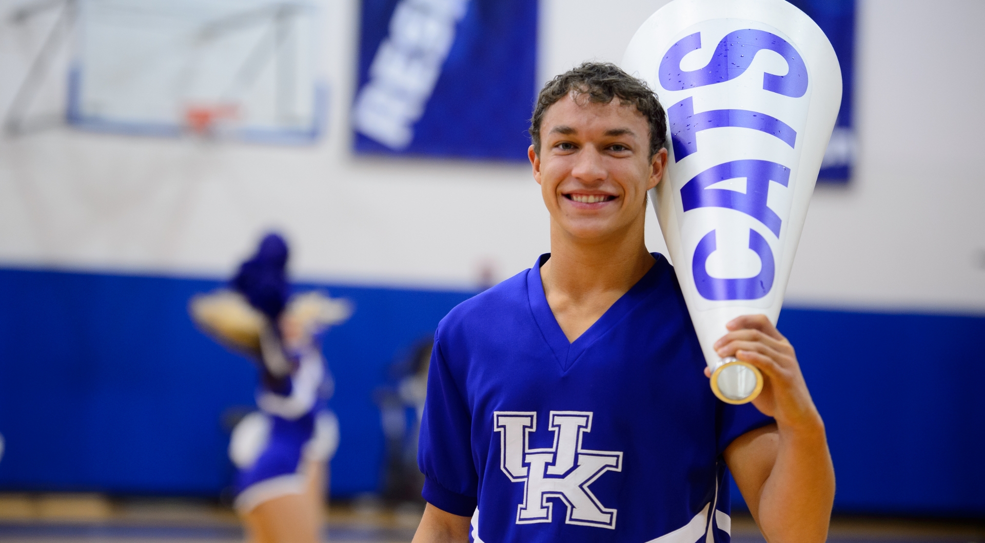 Travis Johnson, a young man with an olive complexion and curly brown hair, smiles for the camera while holding a UK bullhorn. He is wearing a blue and white UK male cheerleader uniform.