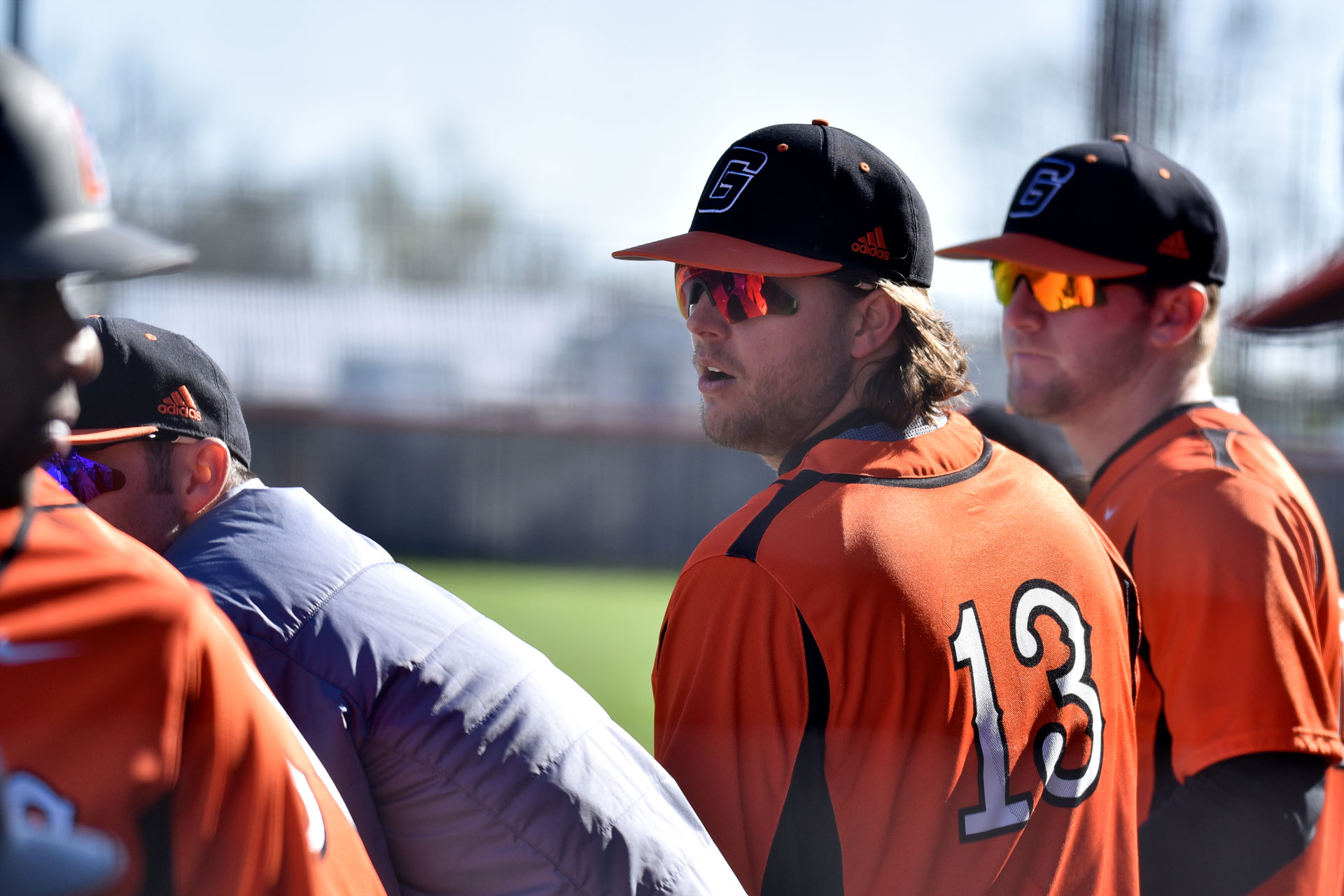 Trent Martin, a college-age white man, stands next to his baseball teammates. He’s wearing an orange baseball uniform with the number 13 on his back. He has long brown hair and is wearing a cap and shades.