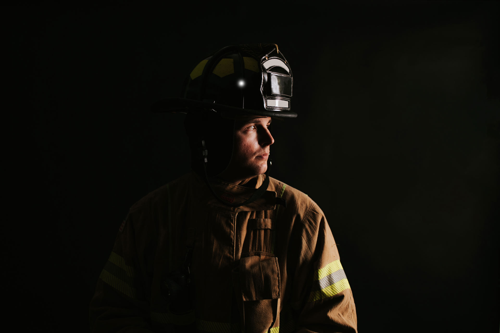 Trent in his firefighting gear and uniform, in front of a black background. His head is turned to the side for a profile view.