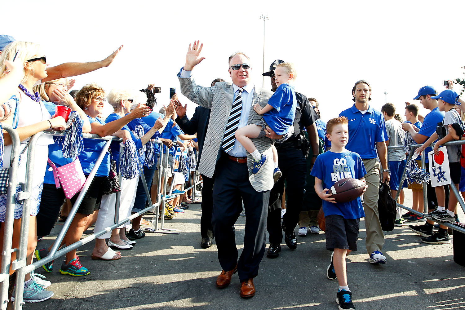 Coach Stoops and his sons walk down the Cat Walk between crowds of fans behind barriers. He is carrying his younger son. HIs older son is carrying a football.